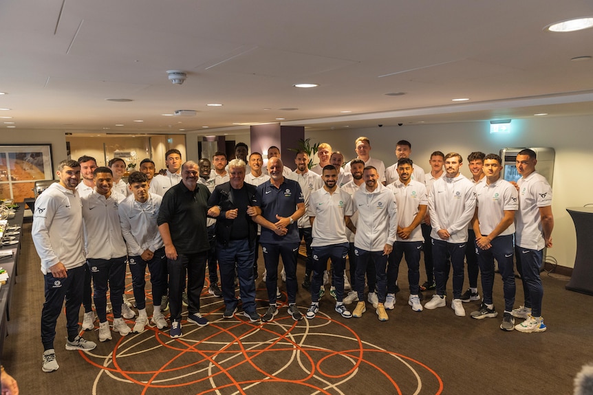 A group of Socceroos players in white tops stand in a room with three current and former Australian coaches.