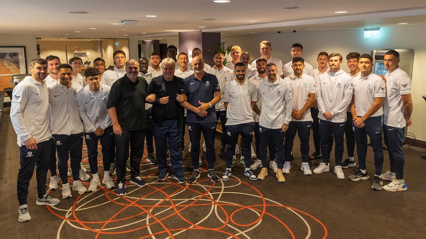 A group of Socceroos players in white tops stand in a room with three current and former Australian coaches.