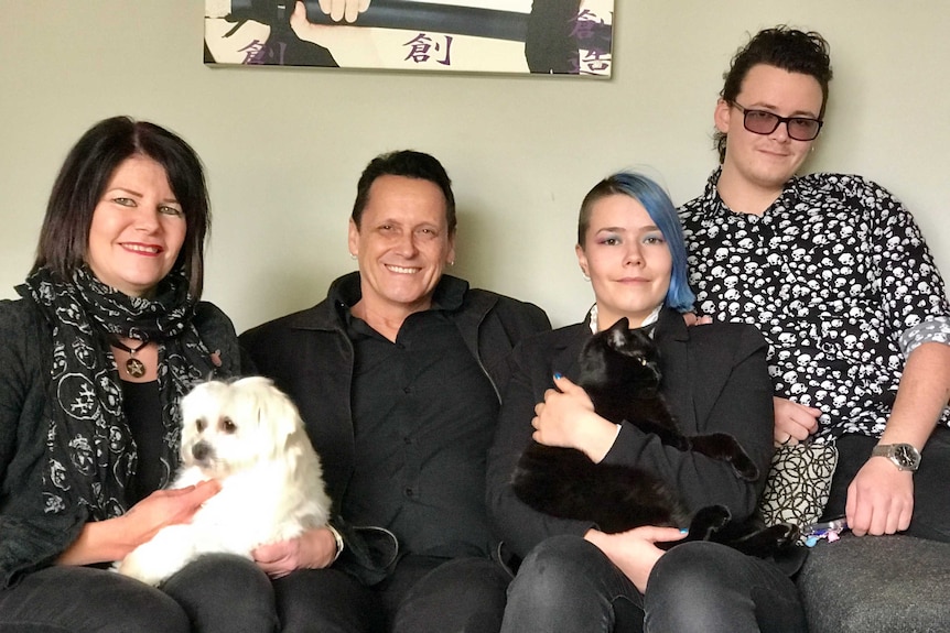 Eden who has blue hair and is holding a black cat sits on a couch with his mother, holding a white lap dog, father, and brother.