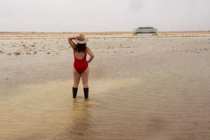 A woman in red swim wear and gum boots stands with her back to the camera on a flooded race track