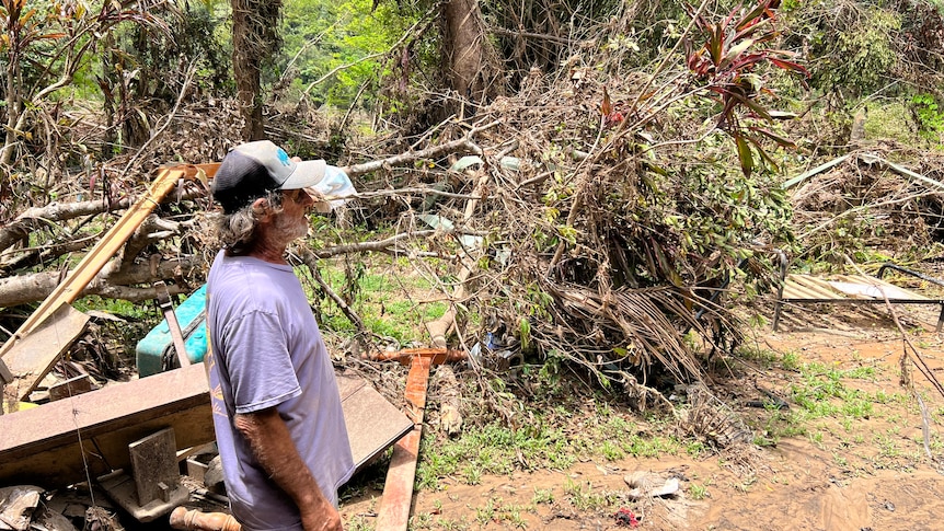 Older man looks out over debris, fallen vegetation and destroyed furniture