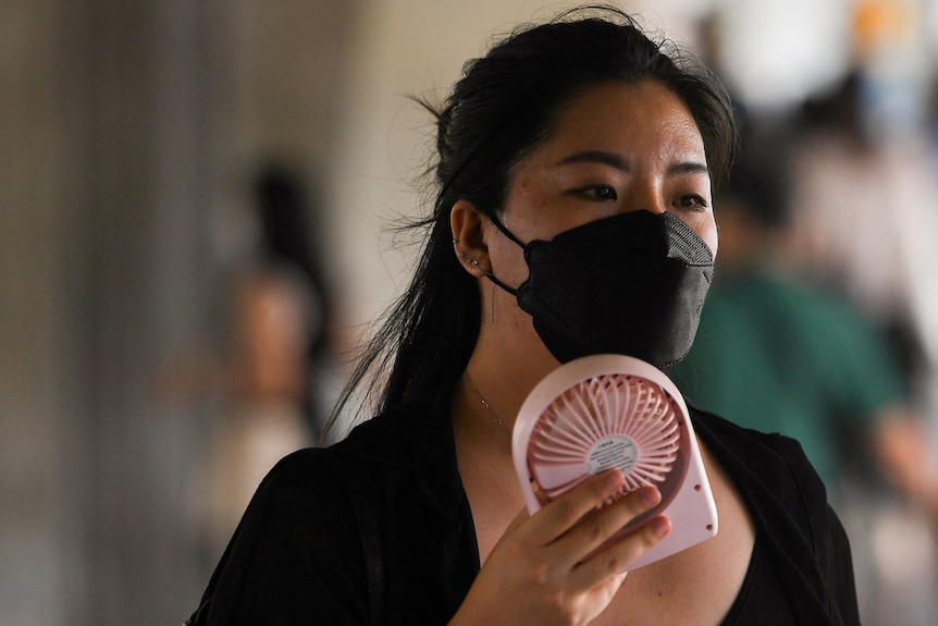 A young Thai woman wearing a dark face mask holds a small pink electric fan up to her face.