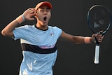 An Australian male professional tennis player holds his right hand to his ear as he celebrates winning a point.
