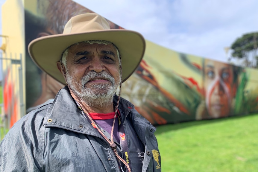 Uncle Dennis wearing a wide-brimmed hat, behind him is a long fence painted with a colourful mural.