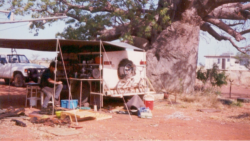 A man sitting under a canopy attached to a trailer under a tree in 1991