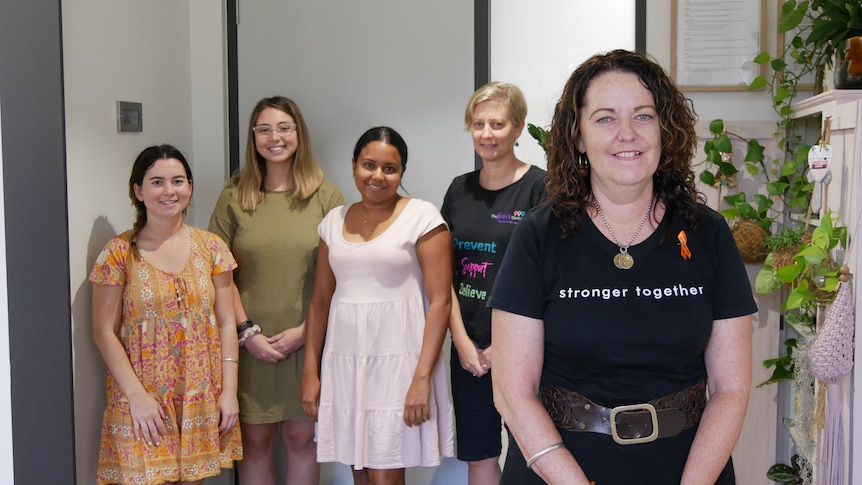 Five women smile at camera in a bright office, next to a bookcase full of plants. 