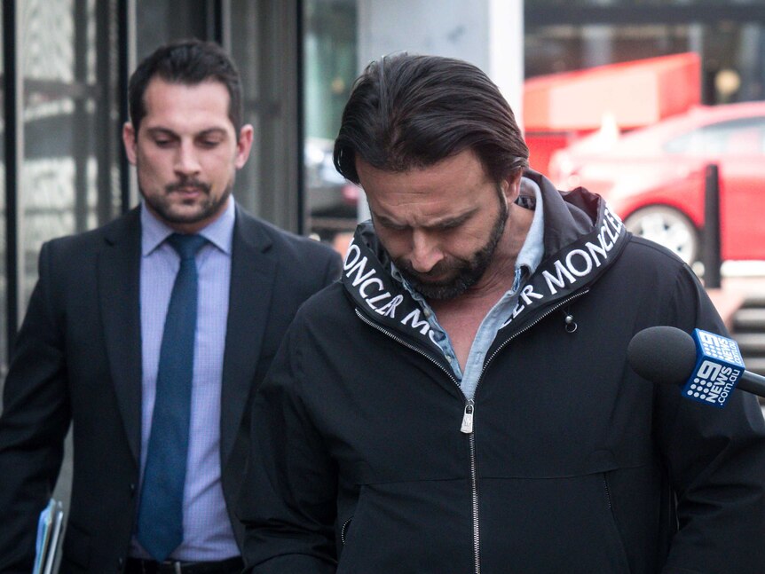 A man wearing a designer jacket walks alongside a male lawyer as members of the media ask him questions.