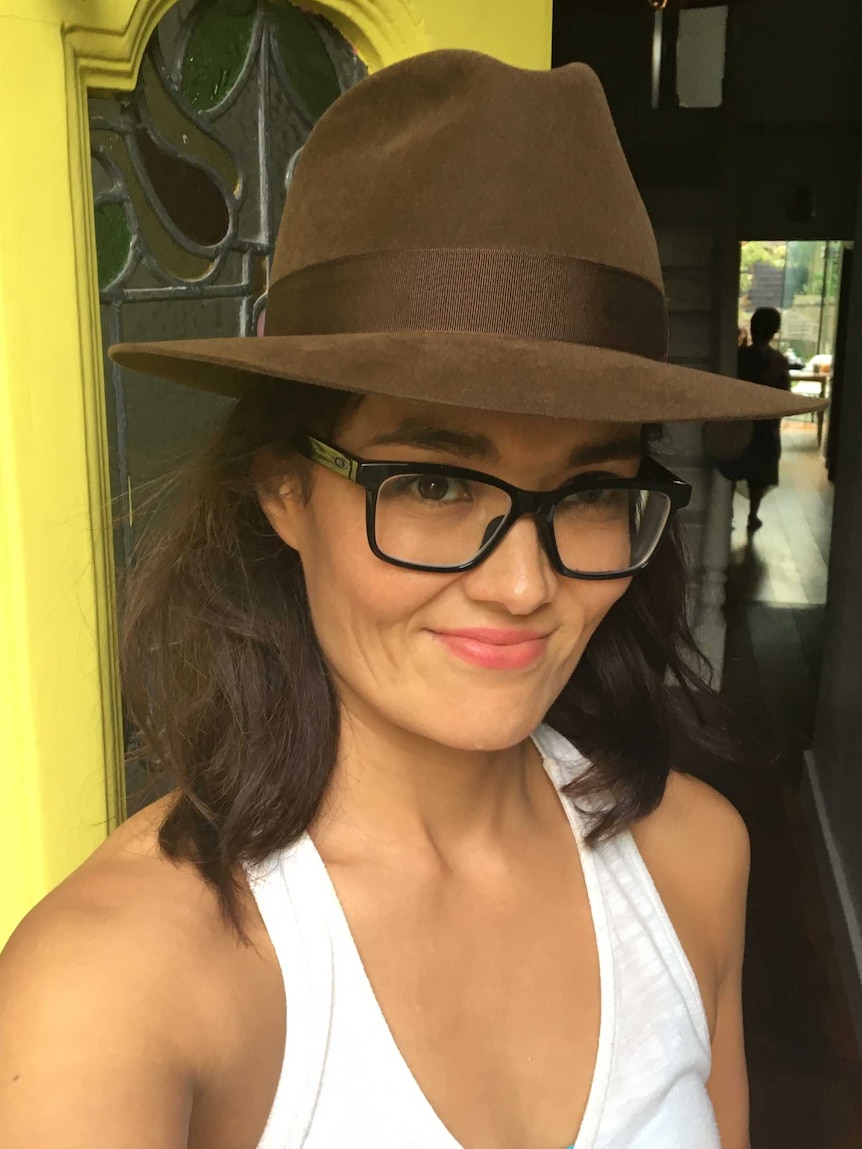 Yumi Stynes stands in front of a yellow door in her favourite brown felt hat
