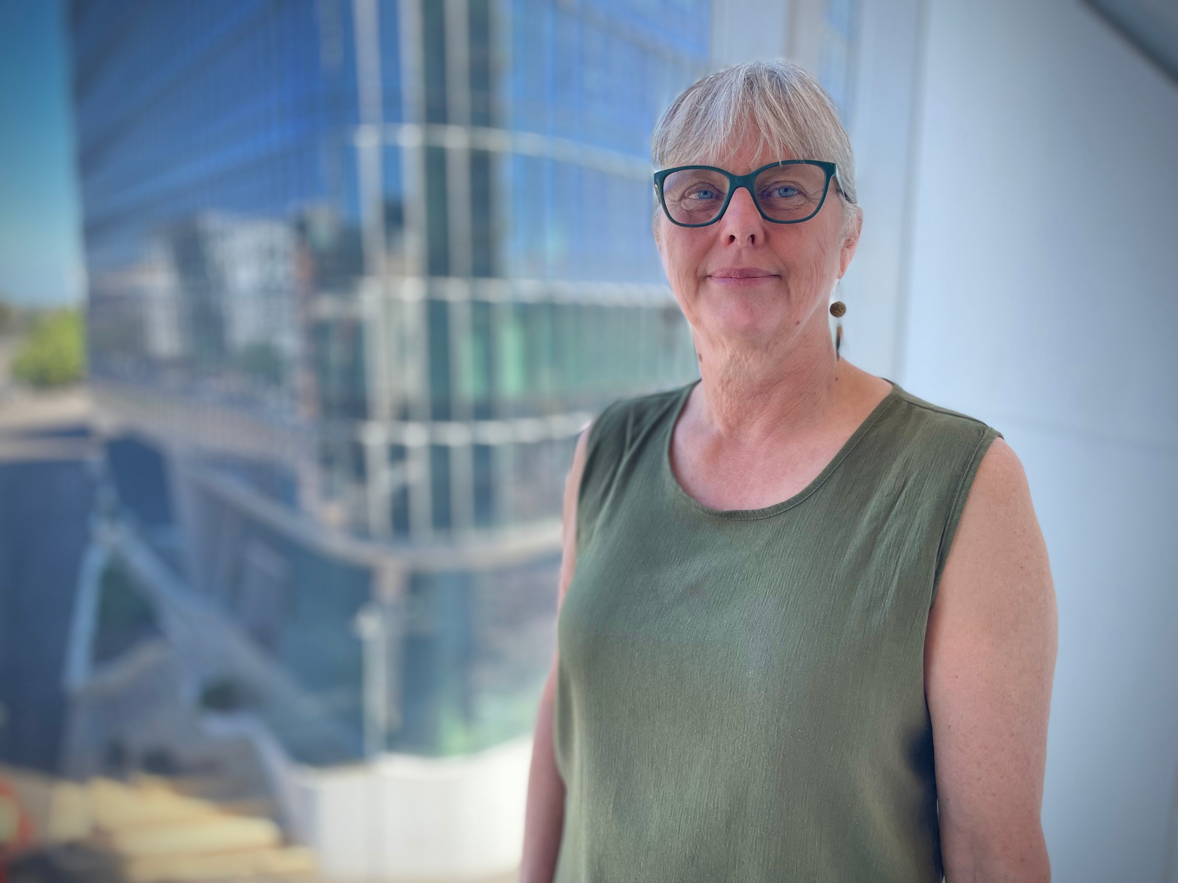A woman wearing glasses and a green shirt standing in front of a window looking out onto a skyline.