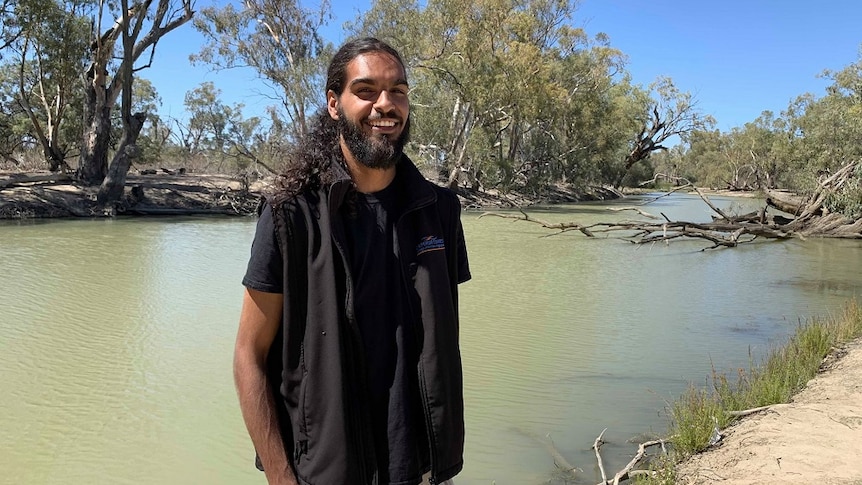 A man is standing on the shore of a lake, smiling. The water is murky and there are native Australian trees behind him