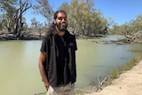 A man is standing on the shore of a lake, smiling. The water is murky and there are native Australian trees behind him