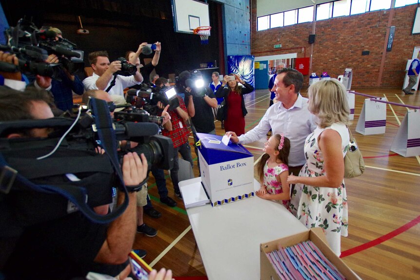 A side shot of Mark McGowan putting a vote in a ballot box with reporters and cameras opposite him.