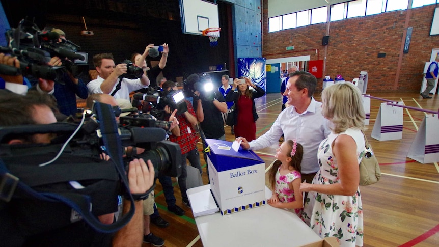 A side shot of Mark McGowan putting a vote in a ballot box with reporters and cameras opposite him.