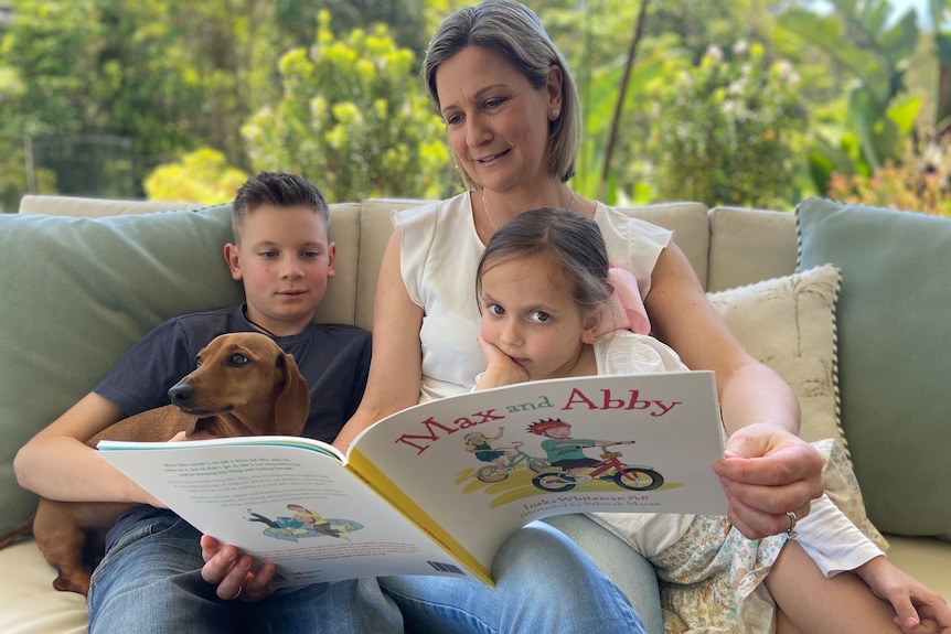 a woman holding a book and sitting on a lounge with two young children