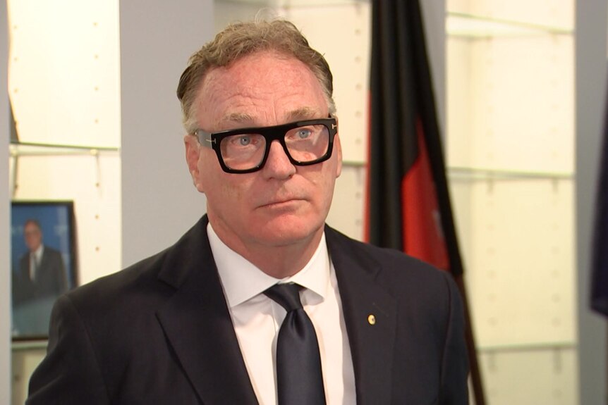 man in thick, black rimmed glasses in suit with Australian and Aboriginal flags in background