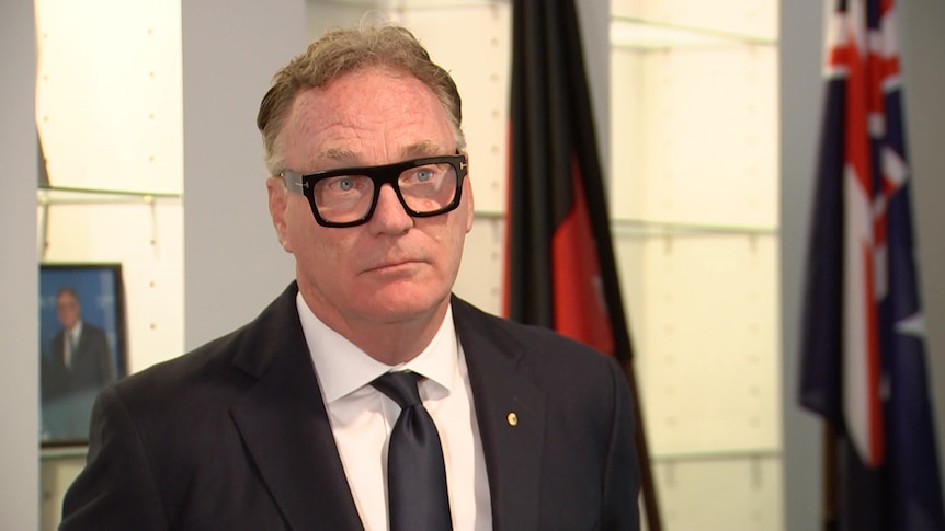 man in thick, black rimmed glasses in suit with Australian and Aboriginal flags in background