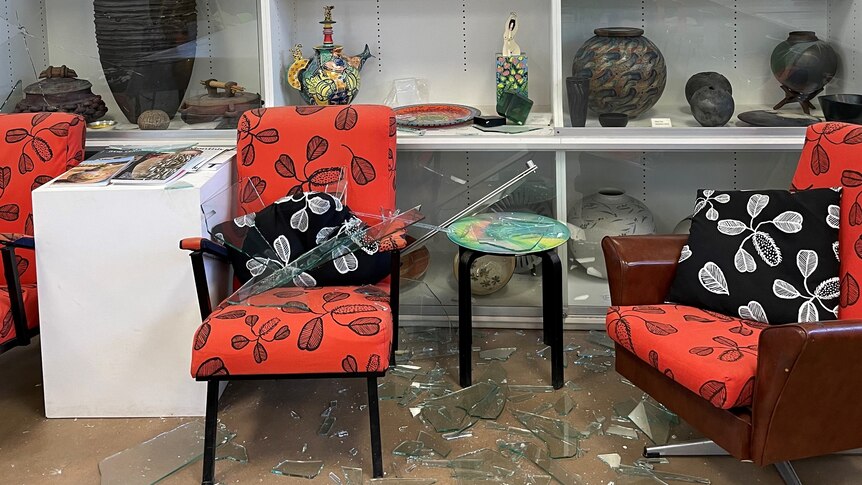 A glass cabinet with art inside is shattered, the shattered glass is on the floor.