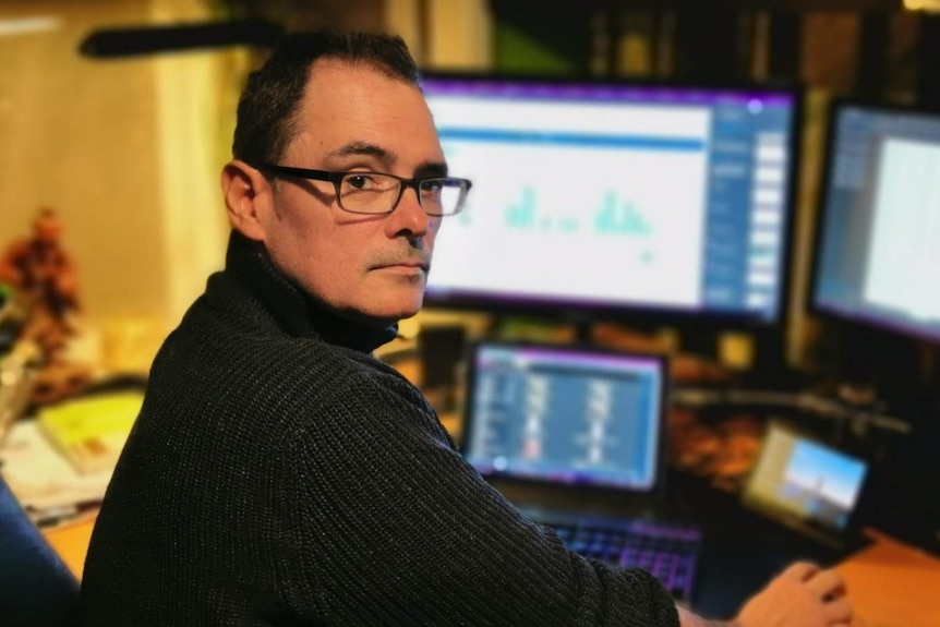 A man wearing glasses looking at the camera in front of computer screens.