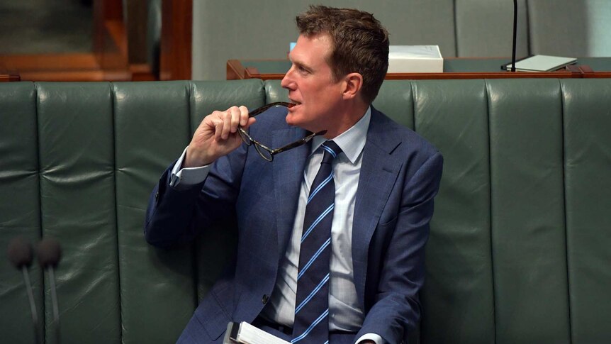Christian Porter sits on the front bench in the House of Representatives, his glasses in his mouth, looking off to his right.