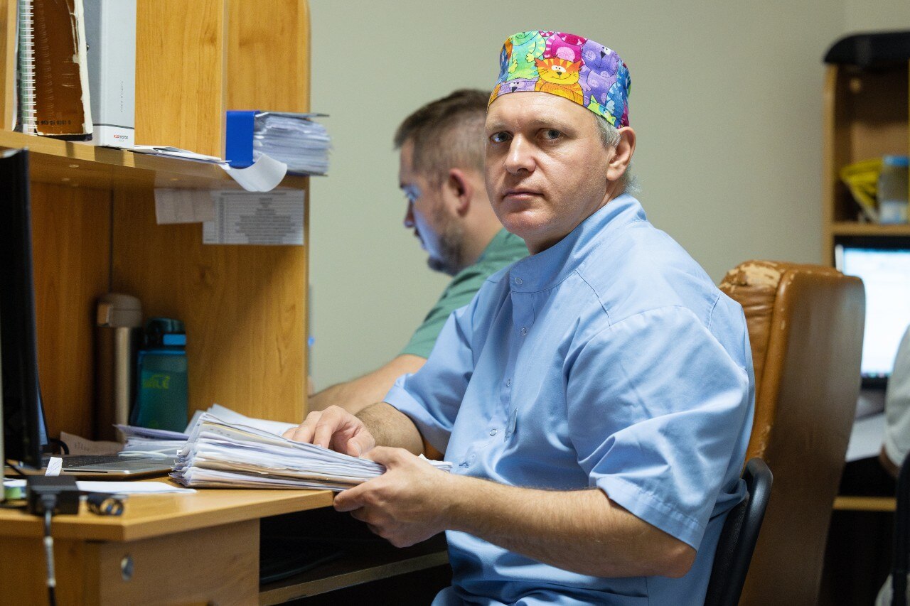 A man with a colourful surgical cap sitting in a chair and looking at the camera with a neutral expression.