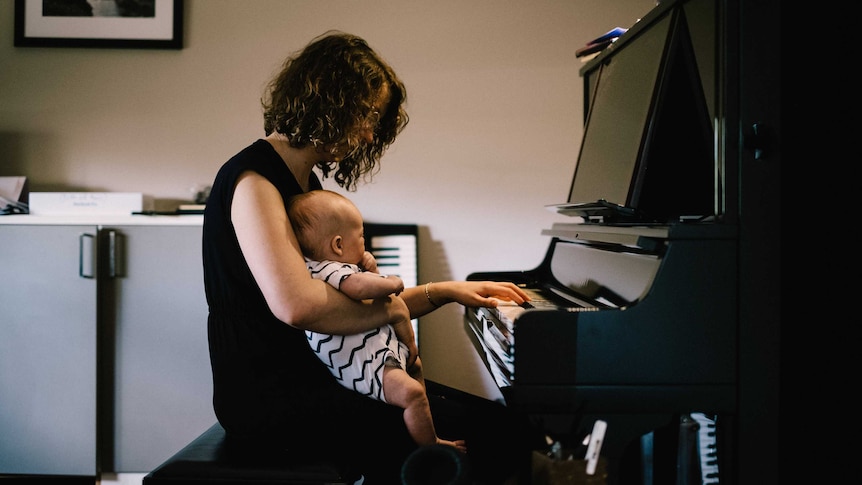 Nat Bartsch sits at an upright piano, playing a chord with her left hand and holding her infant son with her right arm.