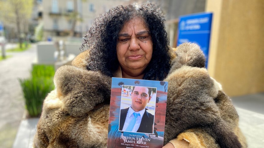 A woman in a possum skin cloak holds a photo of a young man.