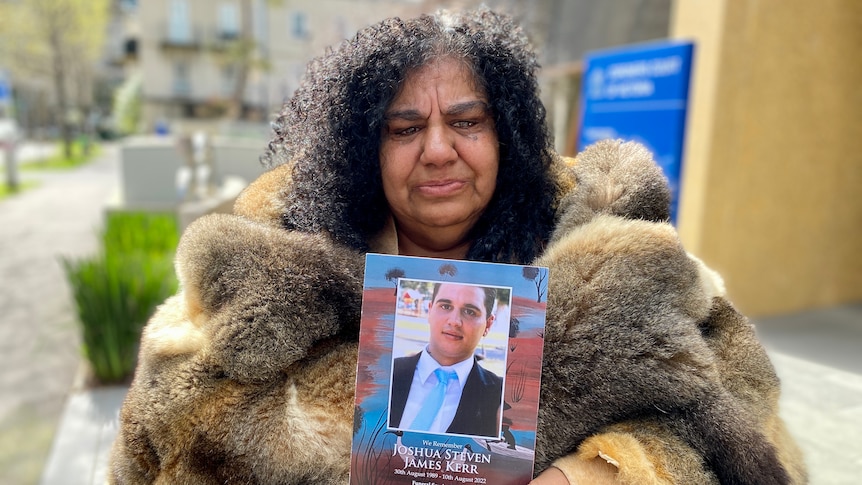 A woman in a possum skin cloak holds a photo of a young man.