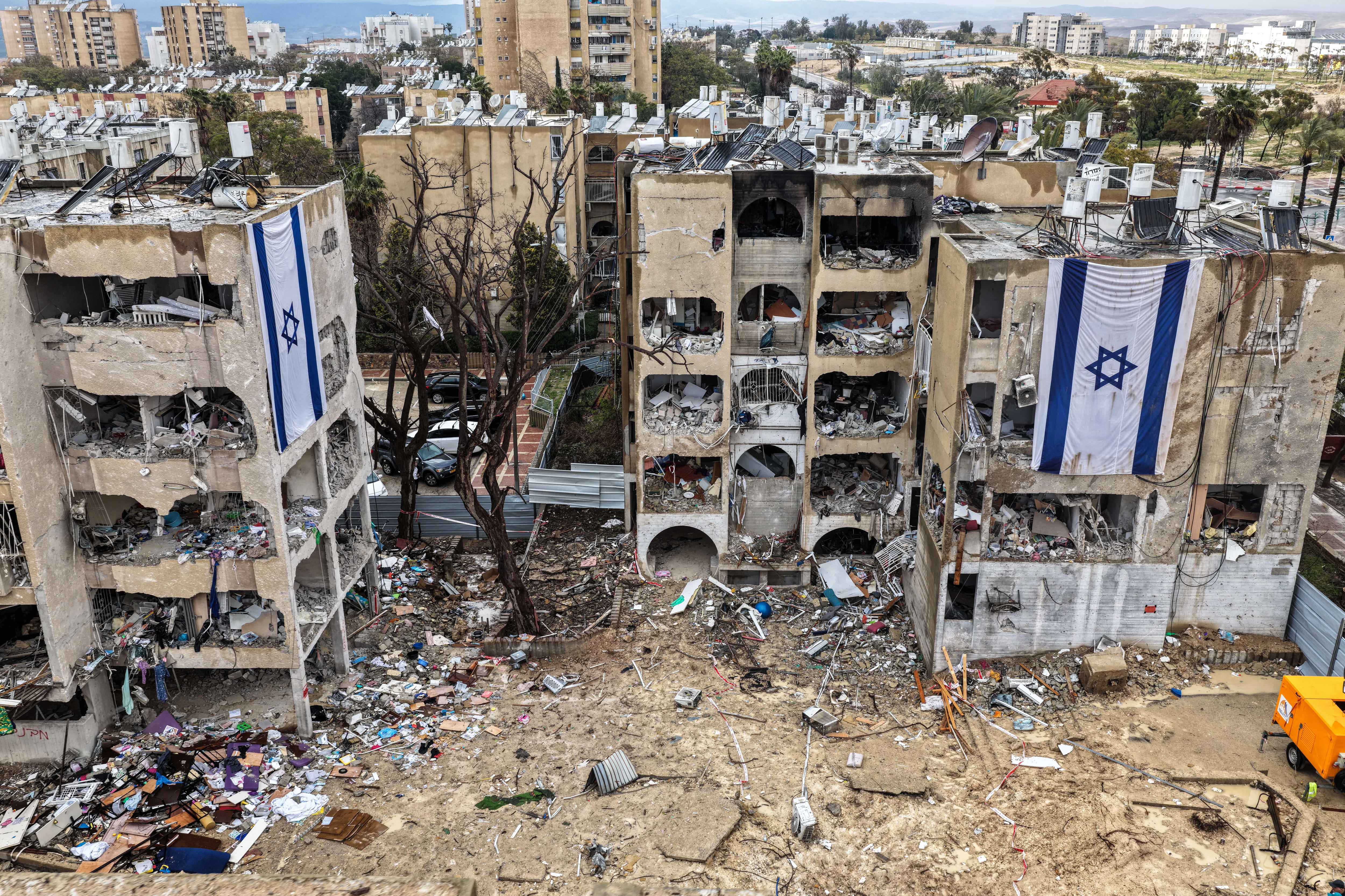 Israeli flags hang from badly damaged, multi-storey buildings in a city. Debris litters the ground around them.