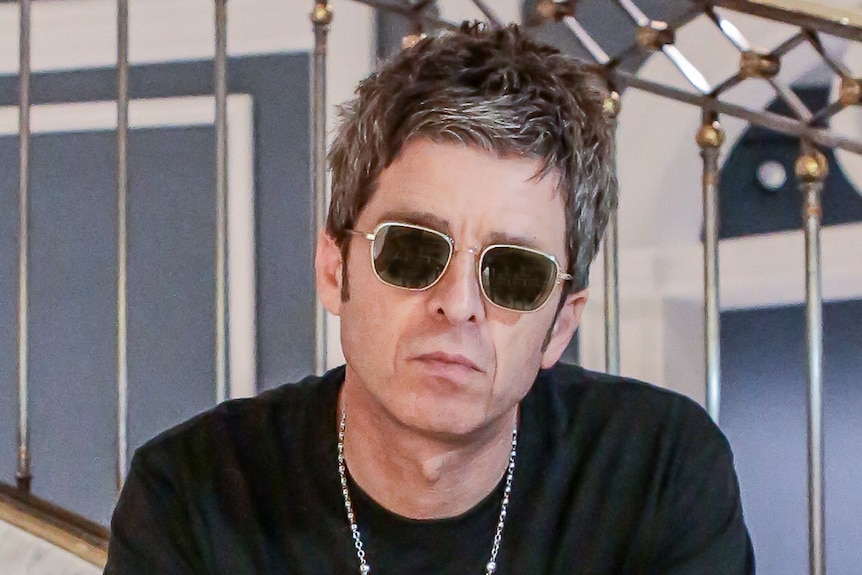 Noel Gallagher glares at the camera wearing sunglasses and a chain necklace