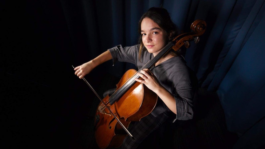 Nina Kiva sits with her cello, looking up at the camera in a dark room