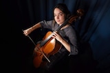Nina Kiva sits with her cello, looking up at the camera in a dark room