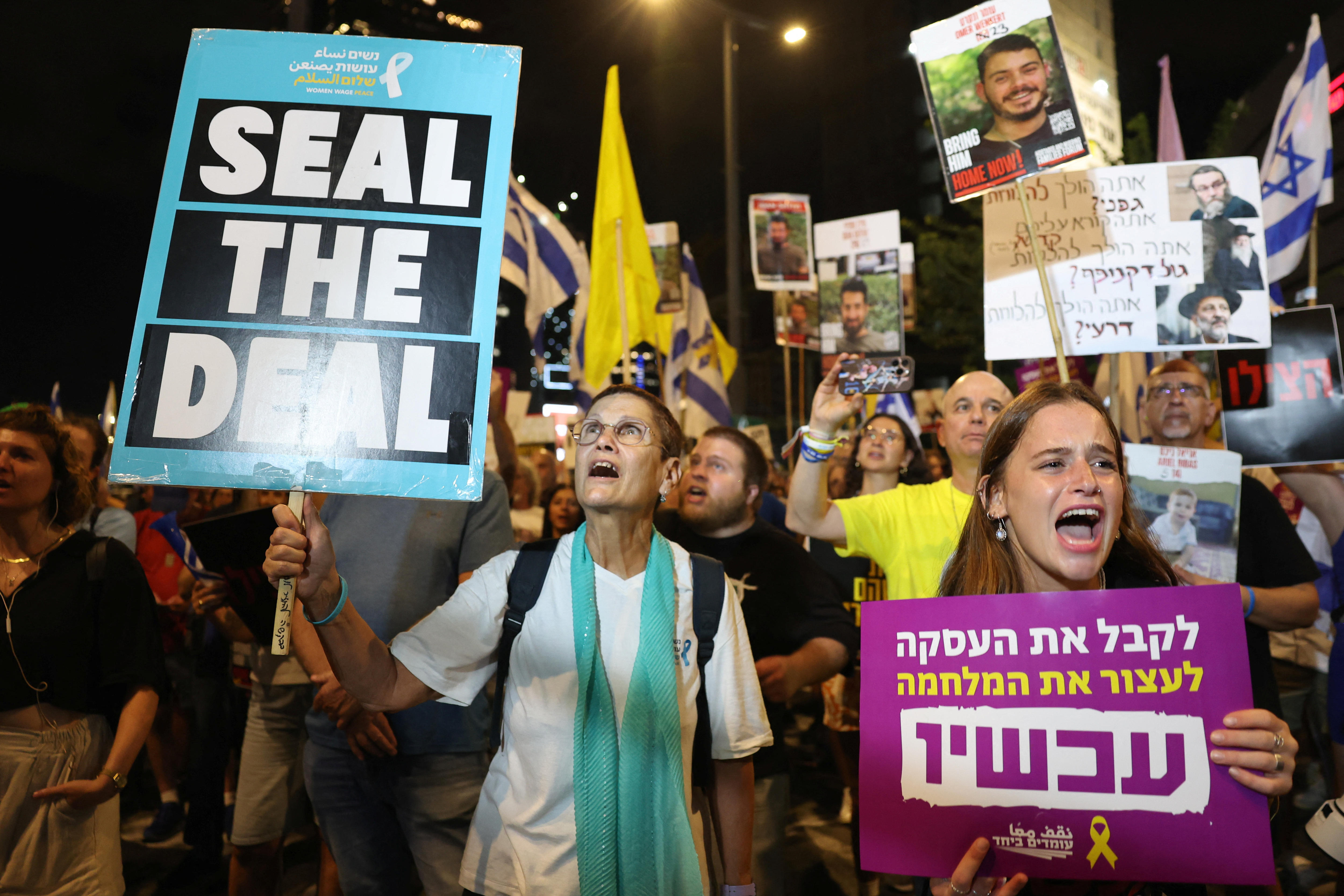 A woman holding up a sign at a protest which reads "Seal the deal" with others around her holding signs with photos on them
