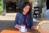 A young Indian woman, straight hair lose, wears denim jacket, striped t-shirts, looks smilingly at phone in hand, drinks coffee.