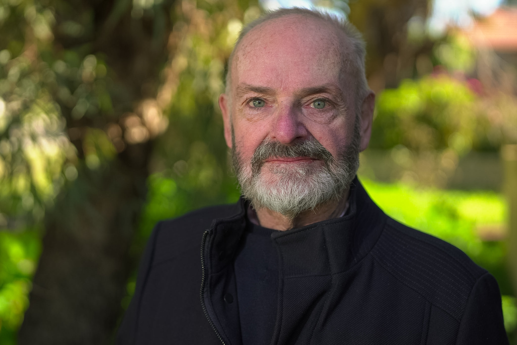 Um homem careca vestindo um casaco sorri para a câmera em frente a um quintal exuberante. 