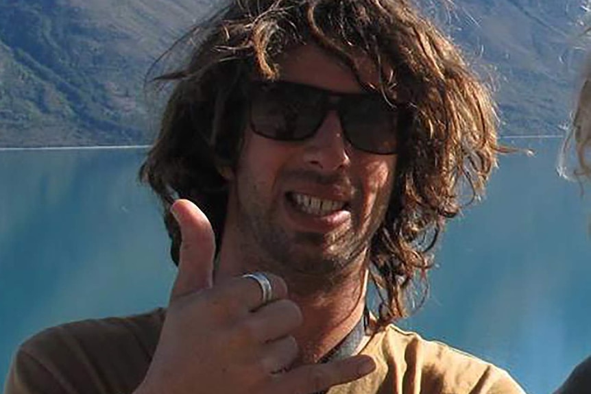 A young man with scruffy brown hair and sunglasses gives the shakas in front of a picturesque mountain and lake