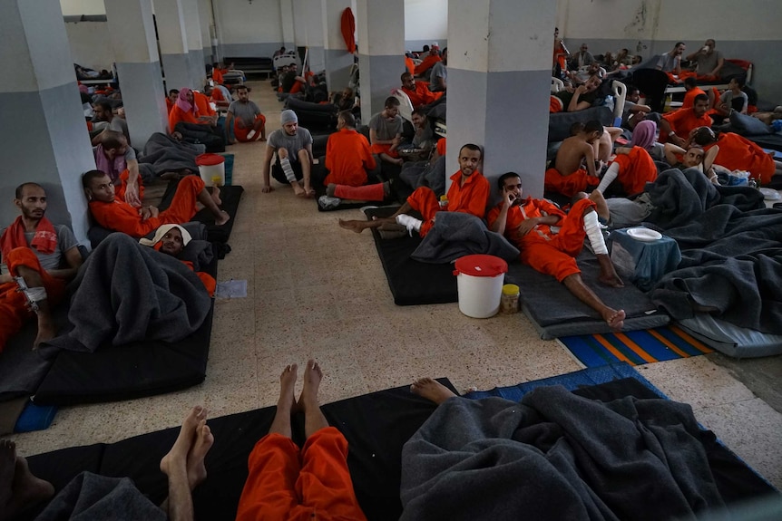 Men in orange prison suits sit down inside a large room.