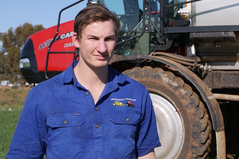 A young man stand in front of large crop spraying machinery, in a field of newly germinating crop.
