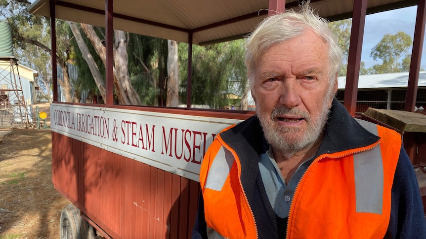 A man wearing an orange jacket standing in front of a wooden cart that says Cobdogla Irrigation and Steam Museum. 