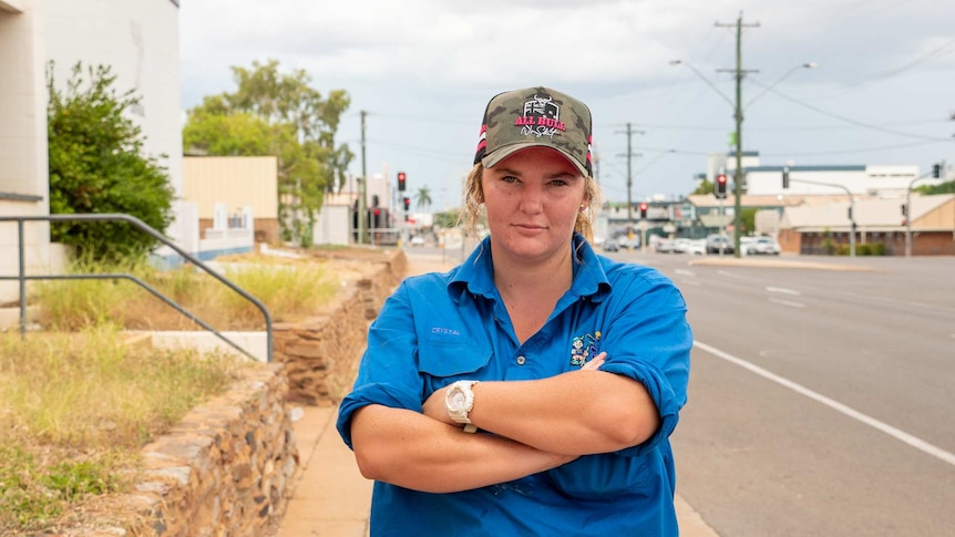 A woman wearing a cap and blue work shirt stands on a footpath, she is not smiling.