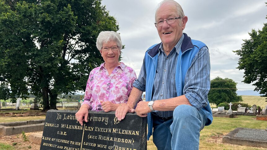  An elderly man and woman stand by a headstone
