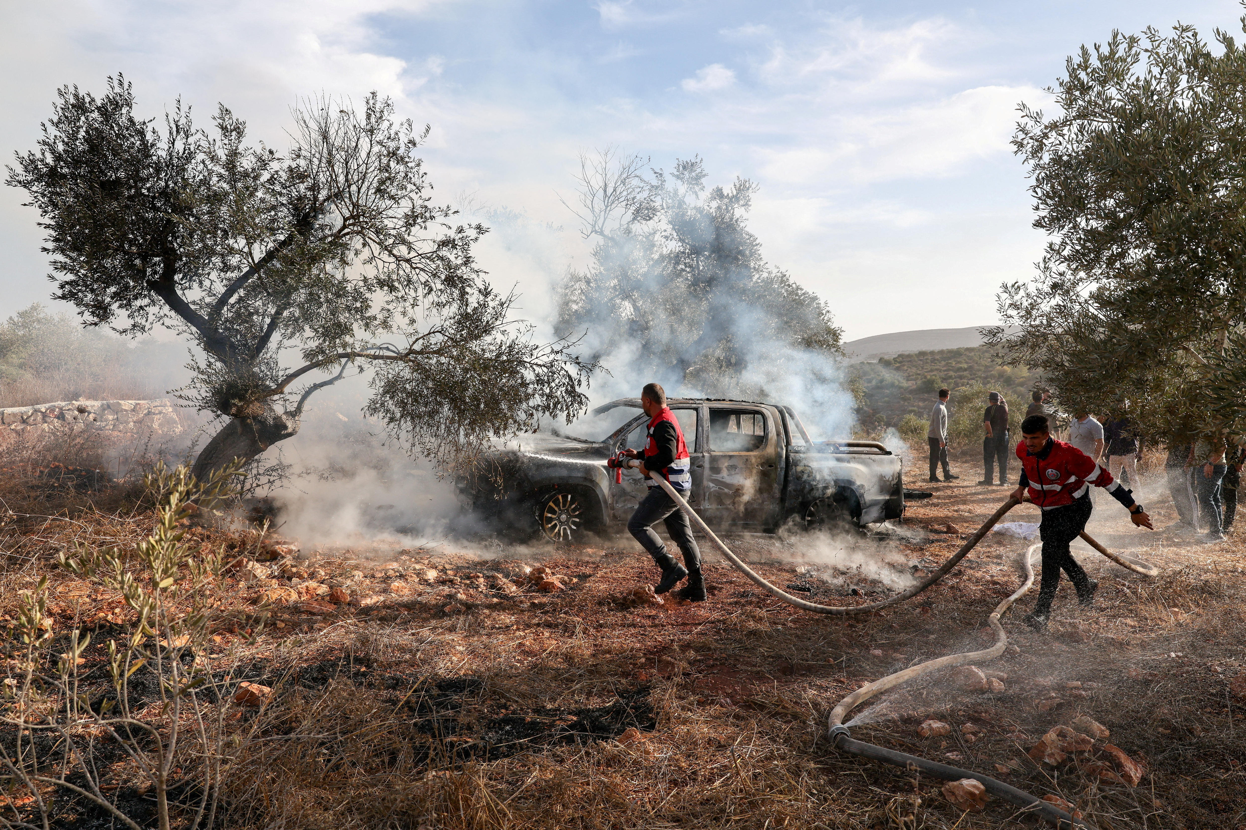 Two firefighters drag a water hose to a smoking, burnt car in an olive grove