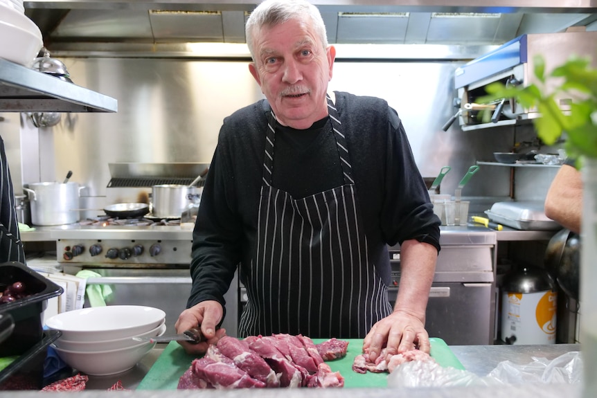 A man with grey hair and moustache stands in a kitchen with a chopping knife in hand.