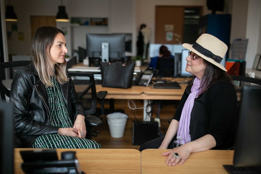 Two women one with a hat face each other in an office