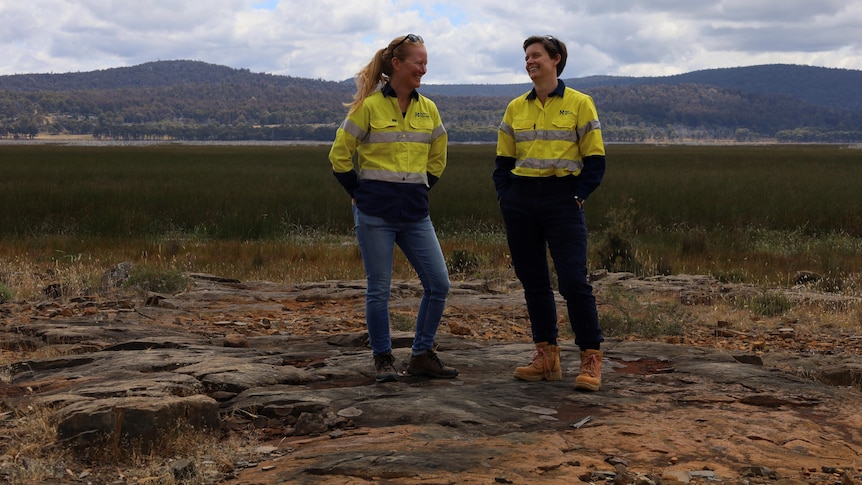 Two women wearing high-vis shirts stand on rocky ground in front of a lagoon filled with reeds