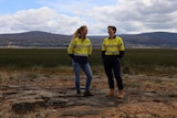 Two women wearing high-vis shirts stand on rocky ground in front of a lagoon filled with reeds