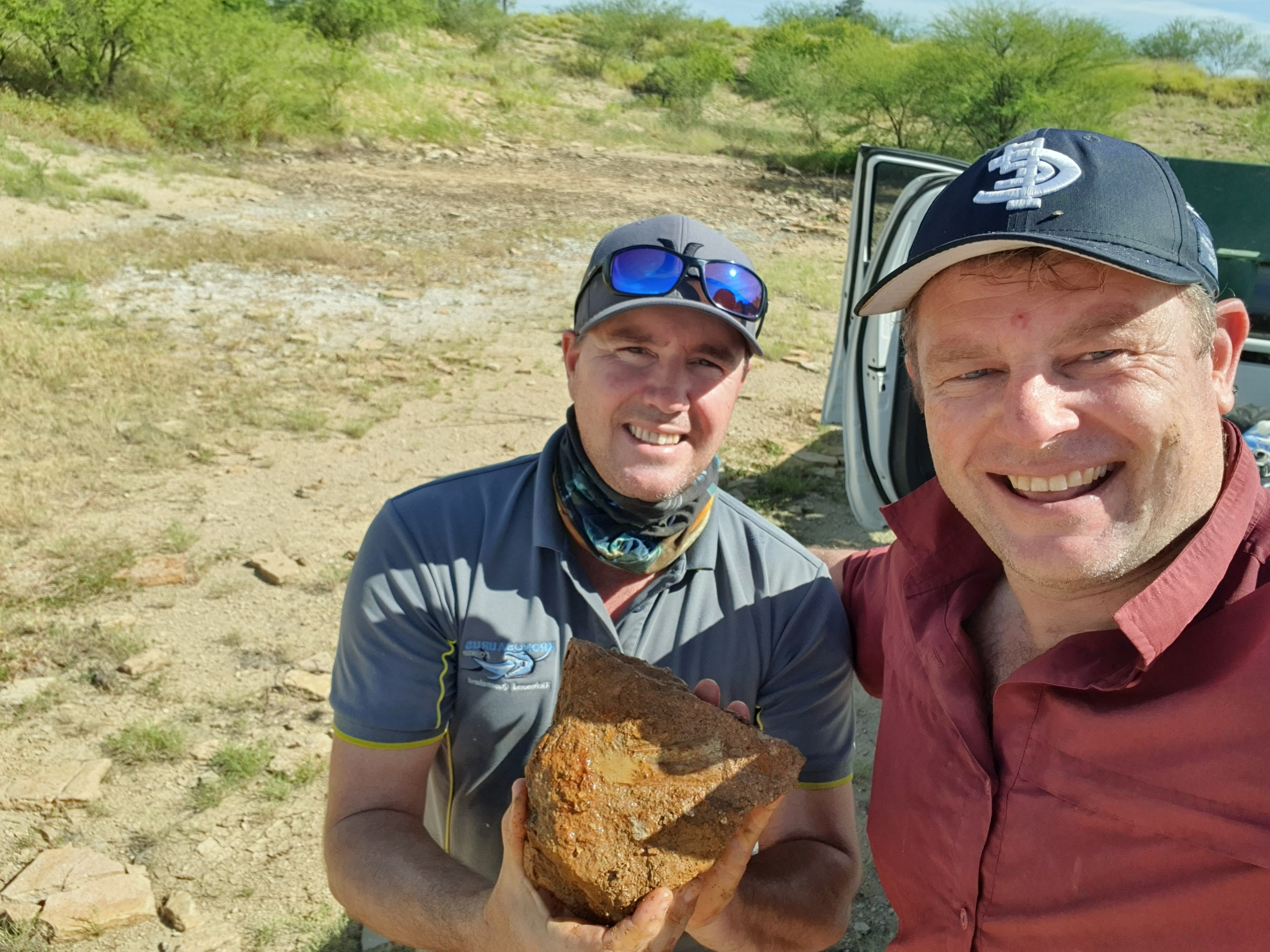 Dois homens sorridentes segurando um fóssil no deserto.