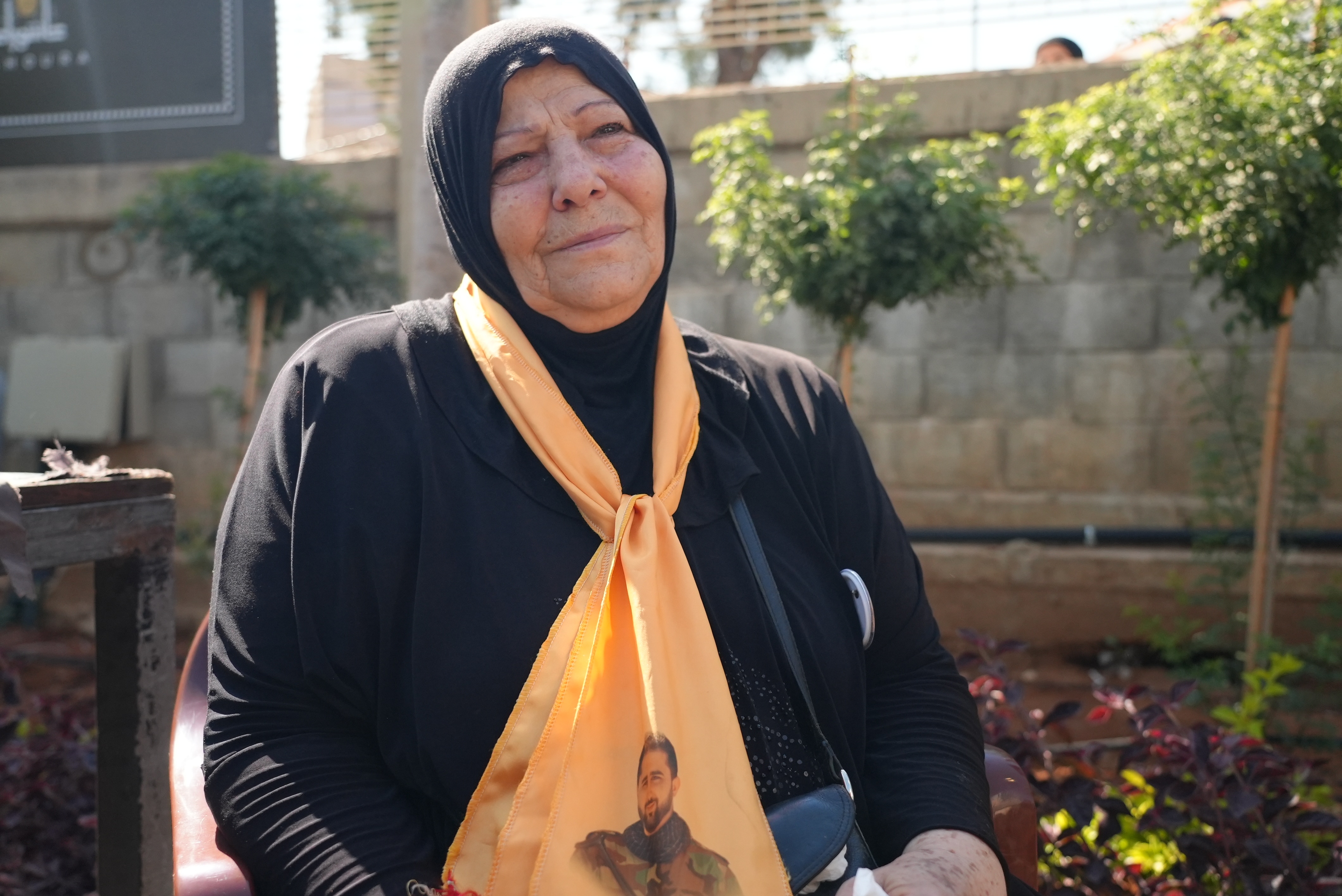 A woman wearing a black hijab and robe. She's also wearing a yellow scarf around her neck with a photo print of her late son.