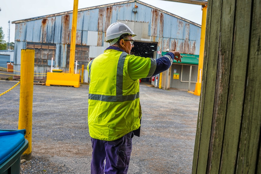 A man in a high vis and hard hat points towards a large workshed.