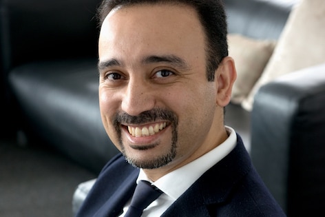 Man in a navy suit with dark hair and moustache smiling sitting on a black leather couch. 
