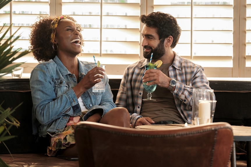 A Black woman and Pakistani American man sit in a bar laughing and drinking